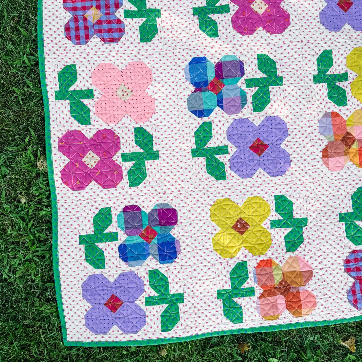 A colorful quilt laid outdoors on green grass, featuring flower and leaf motifs in pink, purple, blue, yellow, green, and red on a pink dotted background.