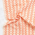 A close-up of draped fabric with a repeating orange and off-white geometric pattern, arranged in soft folds against a light textured background.