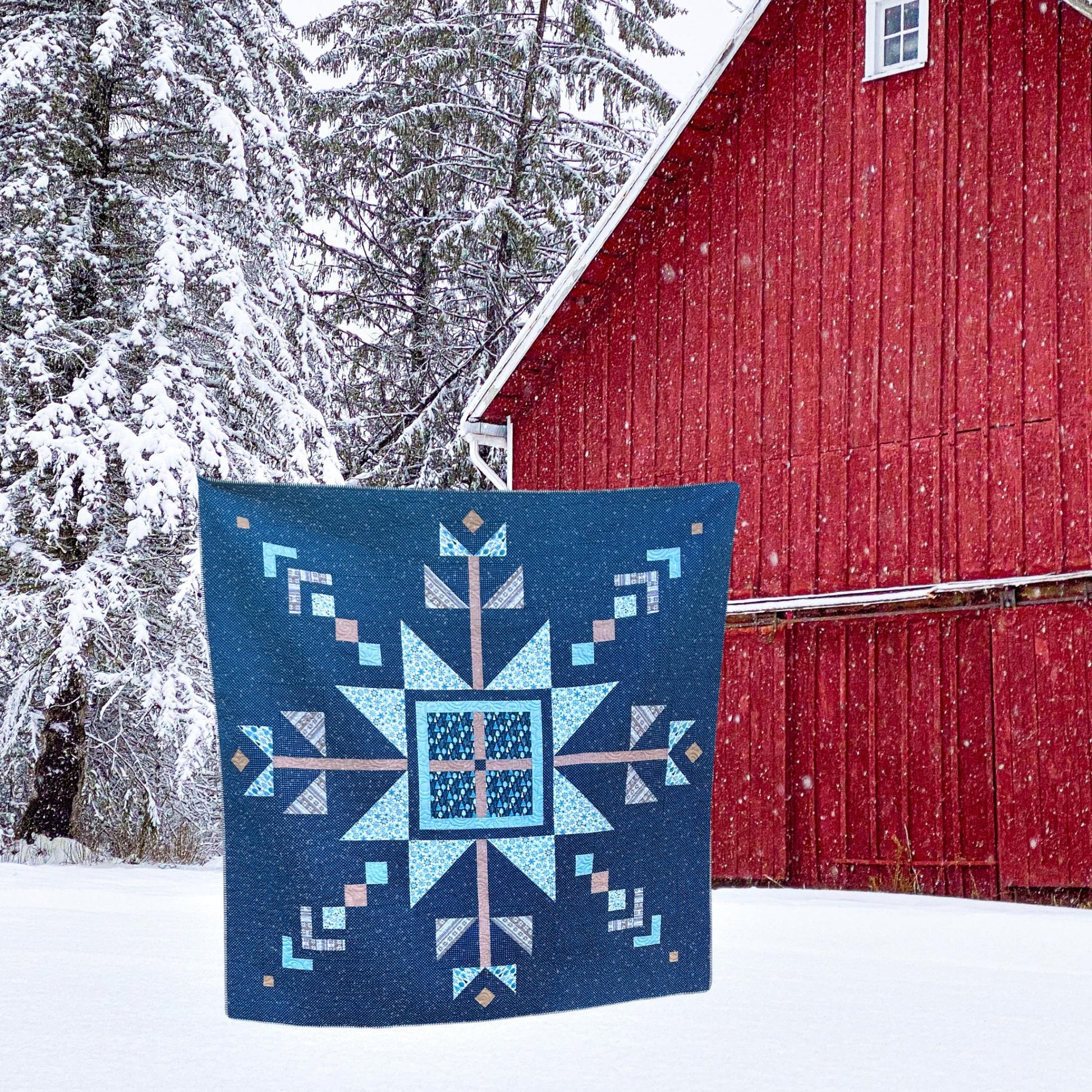 Blue quilt with geometric pattern in front of a red barn during snowfall