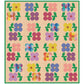 The Mary Golden Quilt quilt pattern by Tamara Darragh for Remi Vail Studio featuring a grid of colorful flower motifs on a warm cream background with  pink dots and a green dot border. The flowers and leaves appear in pink, purple, blue, yellow, orange, and green, with some plaid-style centers.