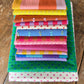 A stack of colorful folded fabrics with various patterns including stripes, polka dots, and floral prints, displayed on a rustic wooden surface.