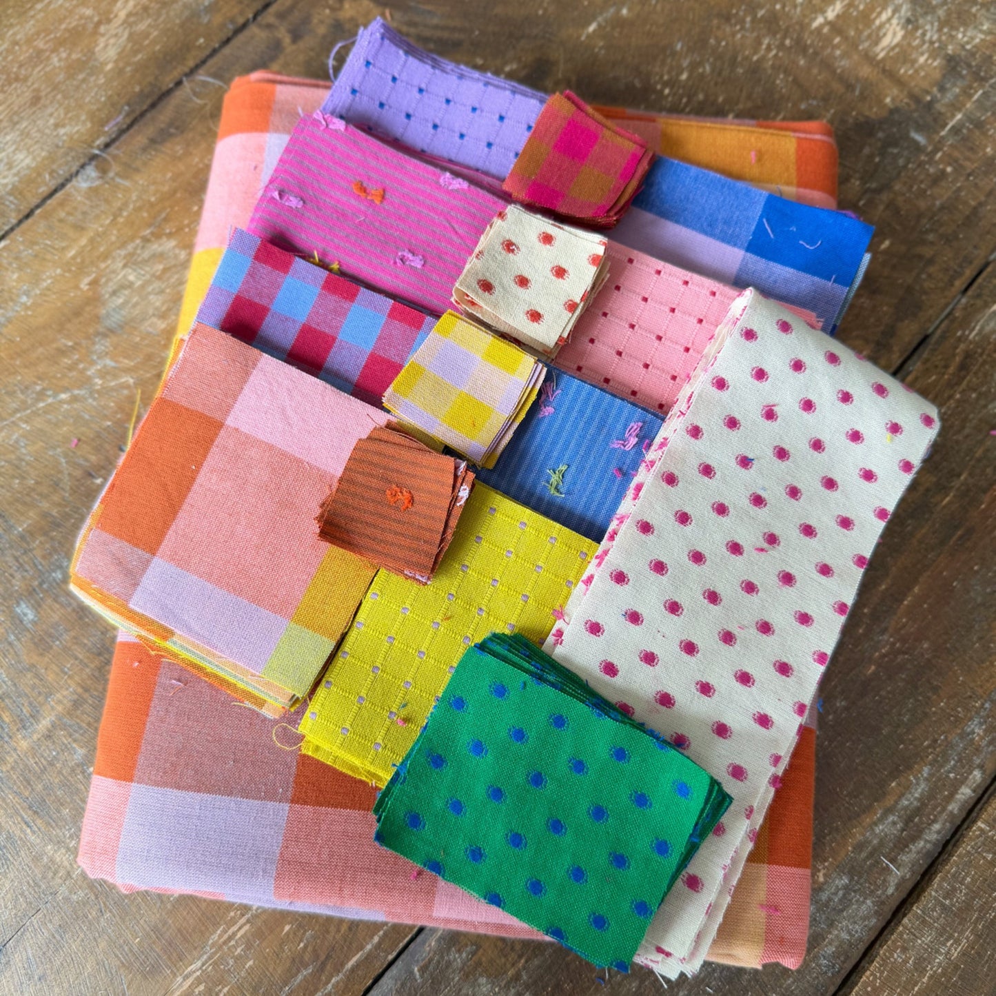 A stack of colorful fabric squares and small folded cloth pieces in assorted patterns including gingham, polka dots, stripes, and checks, arranged on a rustic wooden surface.