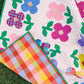 A colorful quilt laid outdoors on grass, featuring large flower patches in pink, blue, red, and purple with green leaves on a pink dotted background. The reverse side is shown, displaying a multicolor plaid pattern with a green binding.