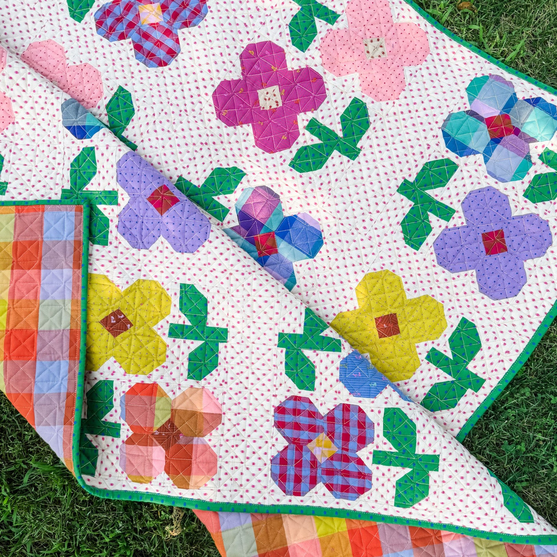 A colorful quilt with a floral patchwork design is laid out on green grass. The quilt features large flowers in pink, purple, yellow, blue, and red gingham patterns on a white background with tiny pink dots, with a green binding, and plaid backing.