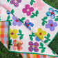 A colorful quilt with a floral patchwork design is laid out on green grass. The quilt features large flowers in pink, purple, yellow, blue, and red gingham patterns on a white background with tiny pink dots, with a green binding, and plaid backing.