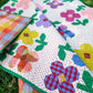 A colorful quilt with a floral patchwork design laid out on green grass. The quilt features large flowers in pink, purple, yellow, blue, and red with green leaves on a white background with small pink polka dots.