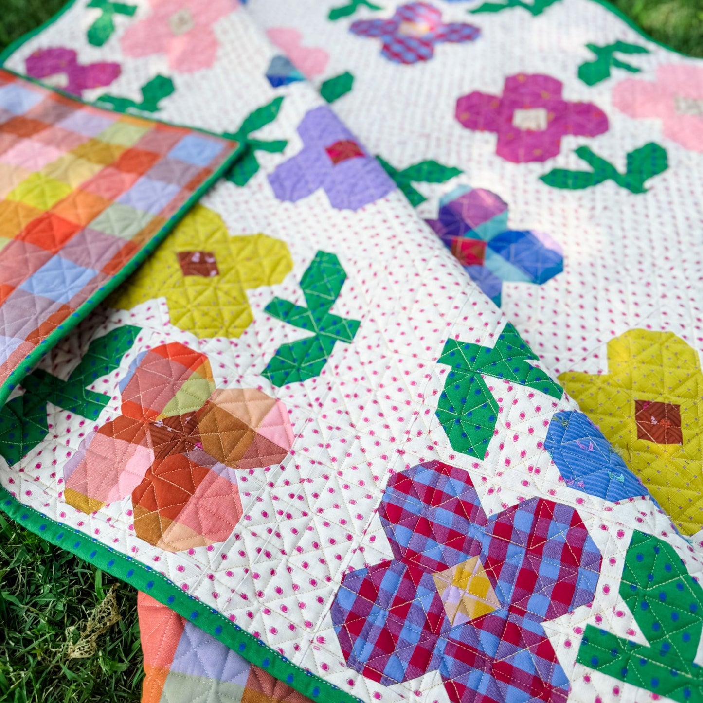 A colorful quilt with a floral patchwork design laid out on green grass. The quilt features large flowers in pink, purple, yellow, blue, and red with green leaves on a white background with small pink polka dots.