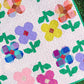 A close-up of a colorful quilt laid outdoors on grass, featuring large flower and leaf motifs in pink, purple, yellow, blue, green, and orange on a white background with small pink dots.