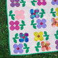 A colorful quilt laid outdoors on green grass, featuring flower and leaf motifs in pink, purple, blue, yellow, green, and red on a pink dotted background.