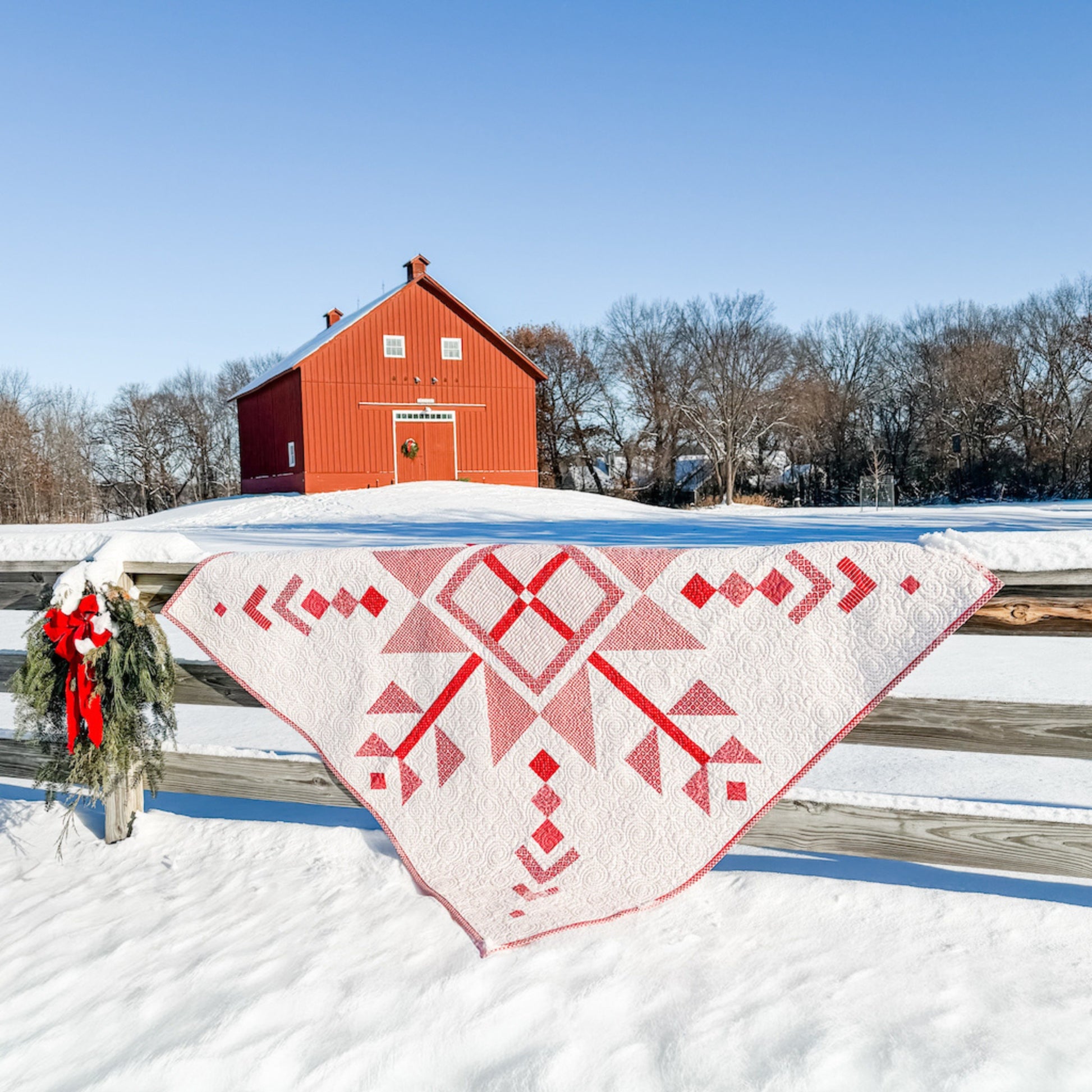 Red and white quilt draped over a wooden fence in front of a red barn in a snowy landscape.