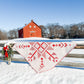 Red and white quilt draped over a wooden fence in front of a red barn in a snowy landscape.