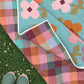 A multicolor Mary Golden Quilt is spread outdoors on grass, with one corner folded back to show the reverse side. The front has large pink flower-like blocks with teal centers and tan accents, while the reverse shows a plaid pattern in pink, orange, purple, teal, and beige.