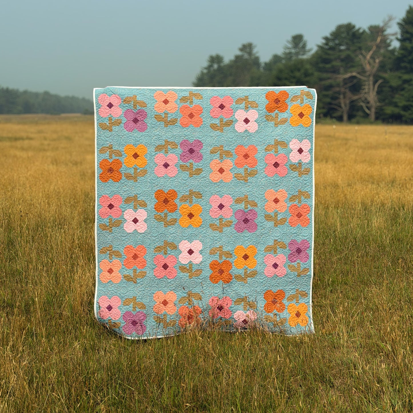 A Limited Palette Mary Golden Quilt by Remi Vail Studio with a patchwork floral pattern in orange, pink, purple, and white on a light blue background, draped in an outdoor grassy field.