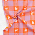 A close-up of a fabric product, featuring a pink and orange gingham check pattern with white woven duck motifs. The fabric is styled with a central twist to show its drape and texture against a light gray background.