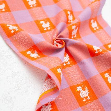 A close-up of a lightweight fabric with a pink and orange gingham check pattern, decorated with small white duck silhouettes, gathered in a swirl on a white textured surface.