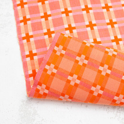 A folded piece of fabric with a plaid pattern in orange, pink, and white, displayed on a light gray textured surface.