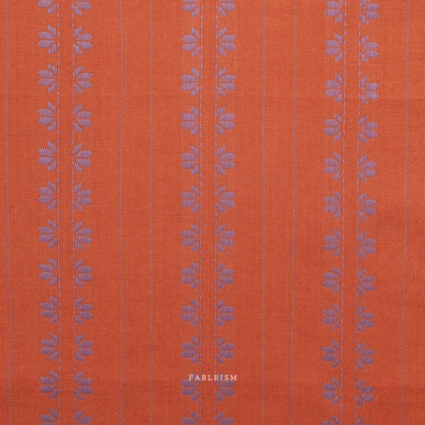 A close-up of textured orange fabric with vertical purple floral motifs and thin purple pinstripes. A small 'FABLEISM' brand name is printed near the bottom center.