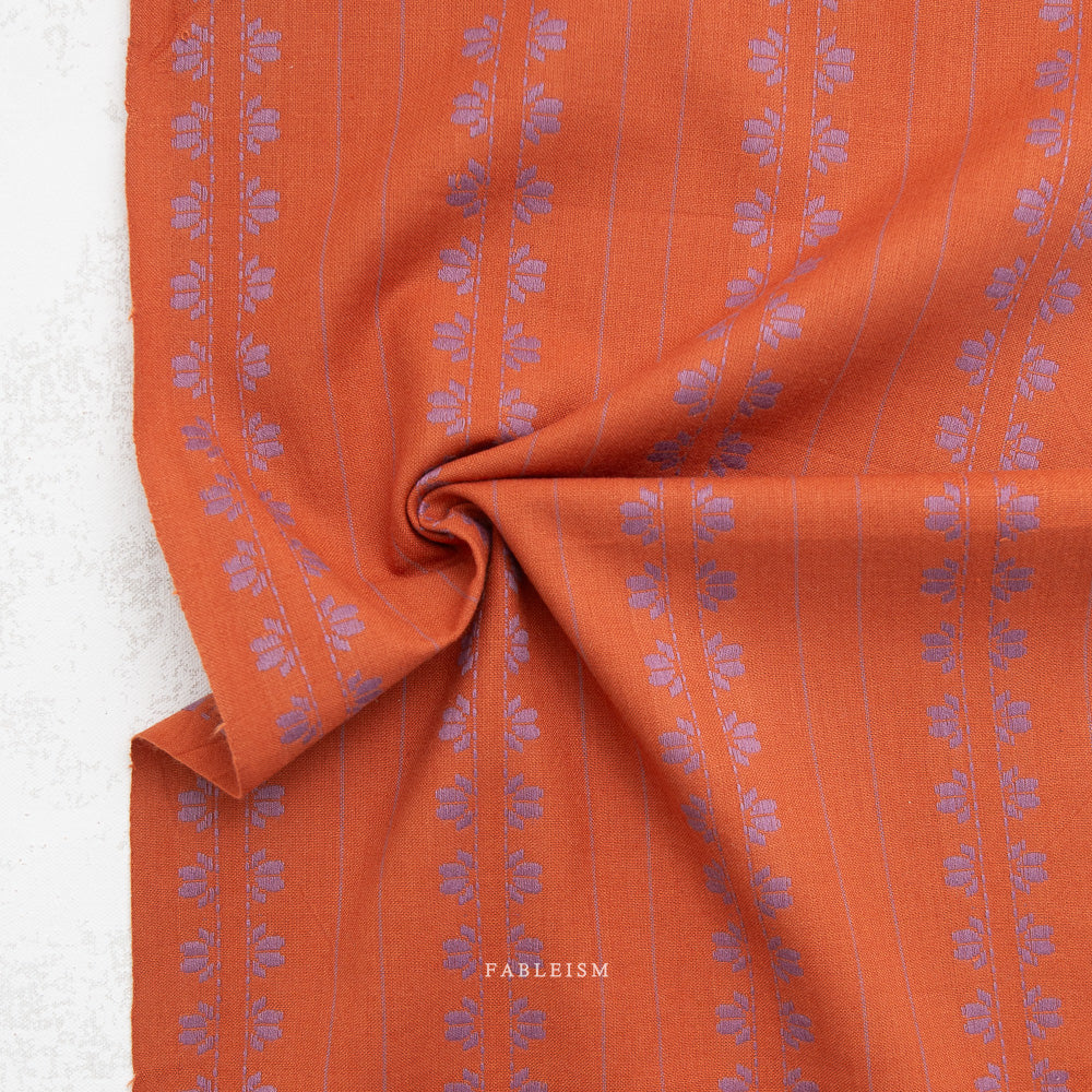 A close-up of orange fabric with a repeating lavender floral pattern and thin vertical stripes, gathered in the center to show texture. The brand name 'FABLEISM' is printed in white near the bottom.