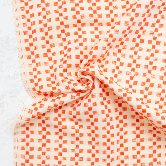 A close-up of draped fabric with a repeating orange and off-white geometric pattern, arranged in soft folds against a light textured background.