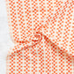 A close-up of draped fabric with a repeating orange and off-white geometric pattern, arranged in soft folds against a light textured background.