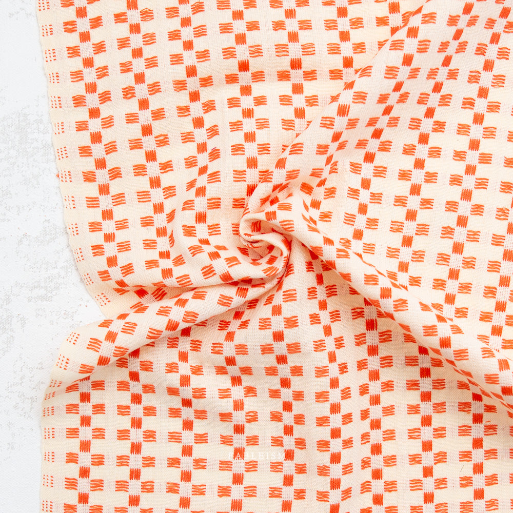 A close-up of draped fabric with a repeating orange and off-white geometric pattern, arranged in soft folds against a light textured background.