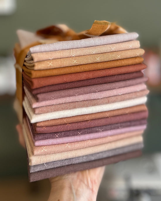 Stack of yarn dyed woven fabrics in shades of peach, brown, pink, cream, and lavender being held up by a hand with a green background.