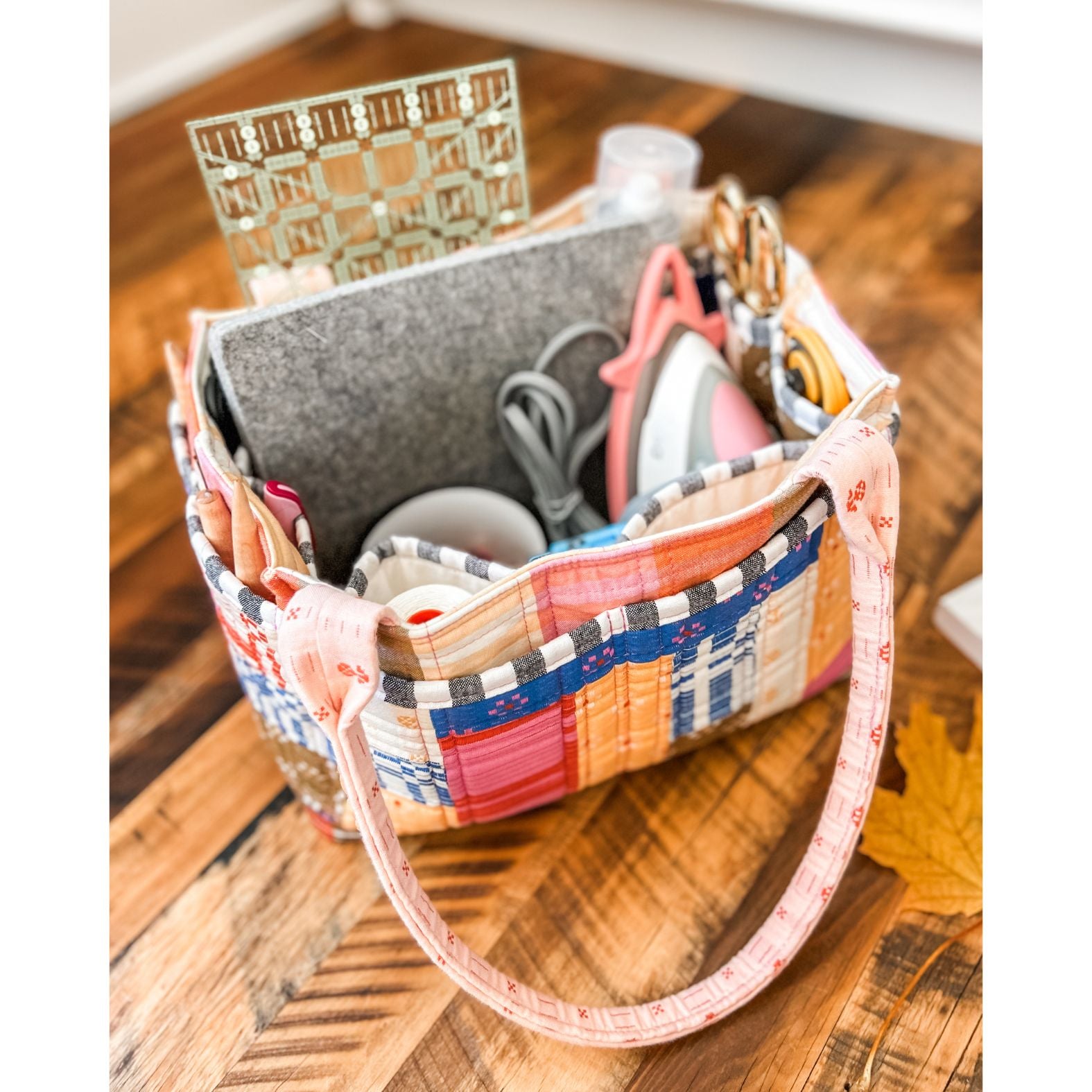 A Patchwork Pockets version of the Maggie Poppins Tote made with yarn dyed wovens in pink, blue, yellow, red, and olive sitting on a wooden floor filled with sewing supplies.
