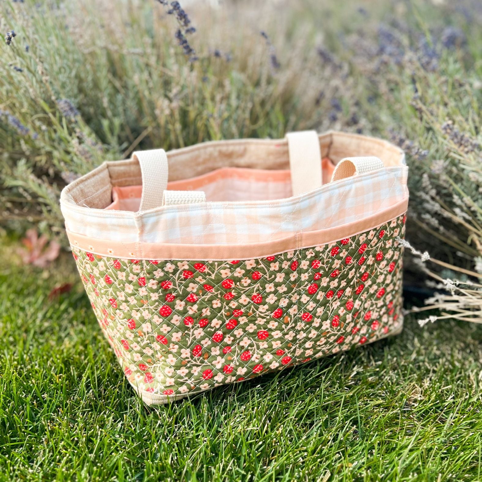 A pink, red, green, and white Maggie Poppins Tote sitting in the grass in front of lavender bushes.