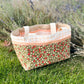 A pink, red, green, and white Maggie Poppins Tote sitting in the grass in front of lavender bushes.
