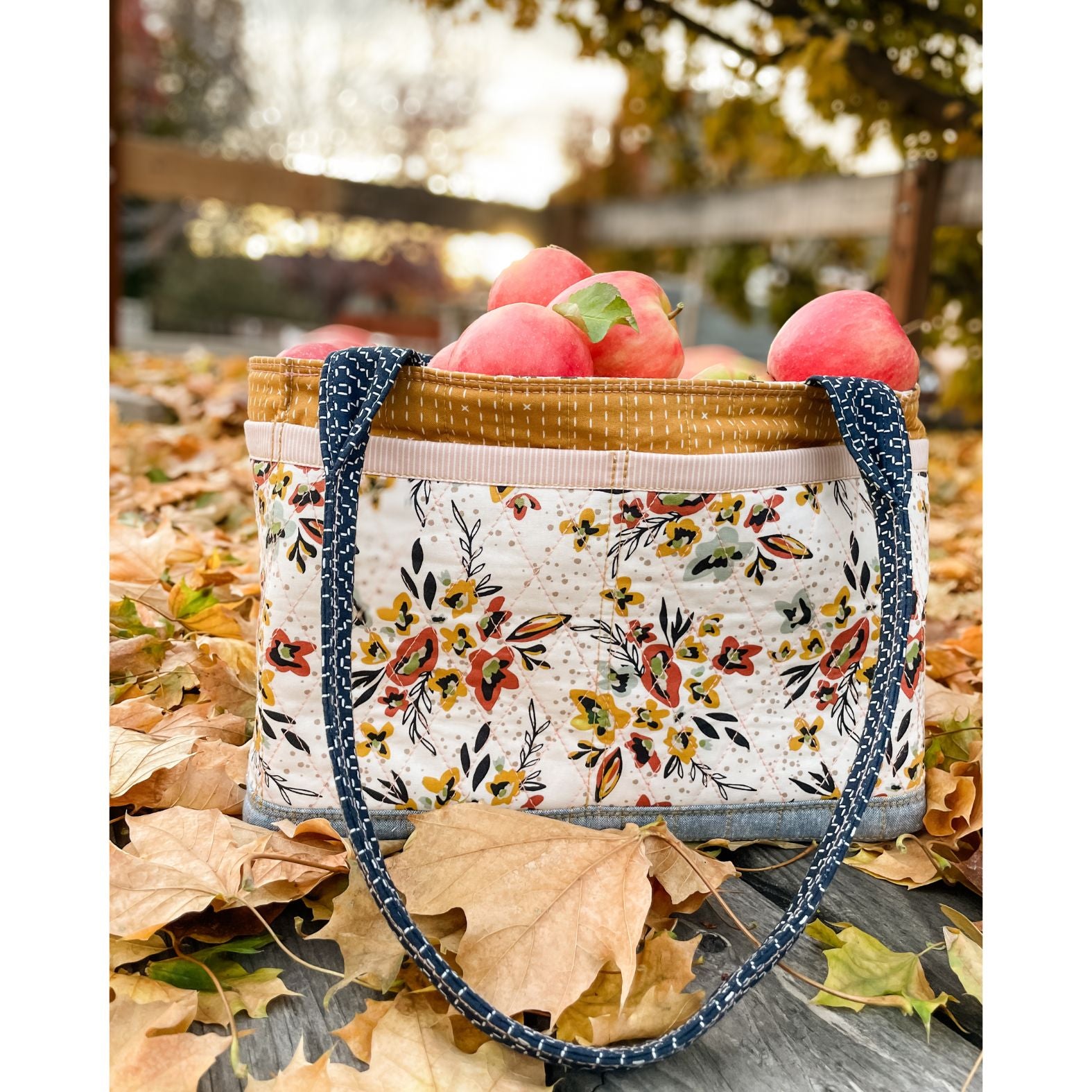 A Maggie Poppins Tote made with pink, brown, rust, white, and black floral fabric filled with apples, sitting on a bed of fallen leaves. 