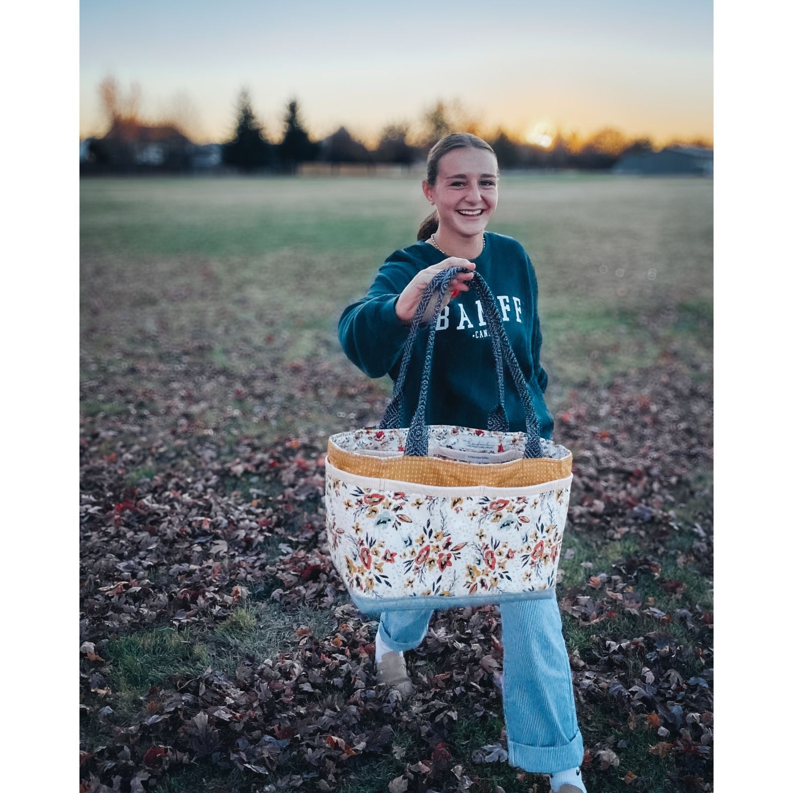 A Maggie Poppins Tote made with pink, brown, rust, white, and black floral fabric held by a smiling girl walking through the leaves and grass at sunset.