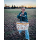 A Maggie Poppins Tote made with pink, brown, rust, white, and black floral fabric held by a smiling girl walking through the leaves and grass at sunset.