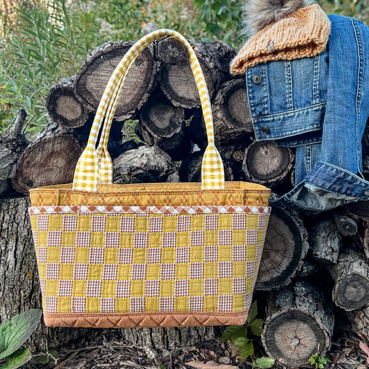 Quilted Maggie Poppns Tote in shades of yellow, brown, and white hanging on a wood pile in a garden with a denim jacket and yellow knit hat next to it.