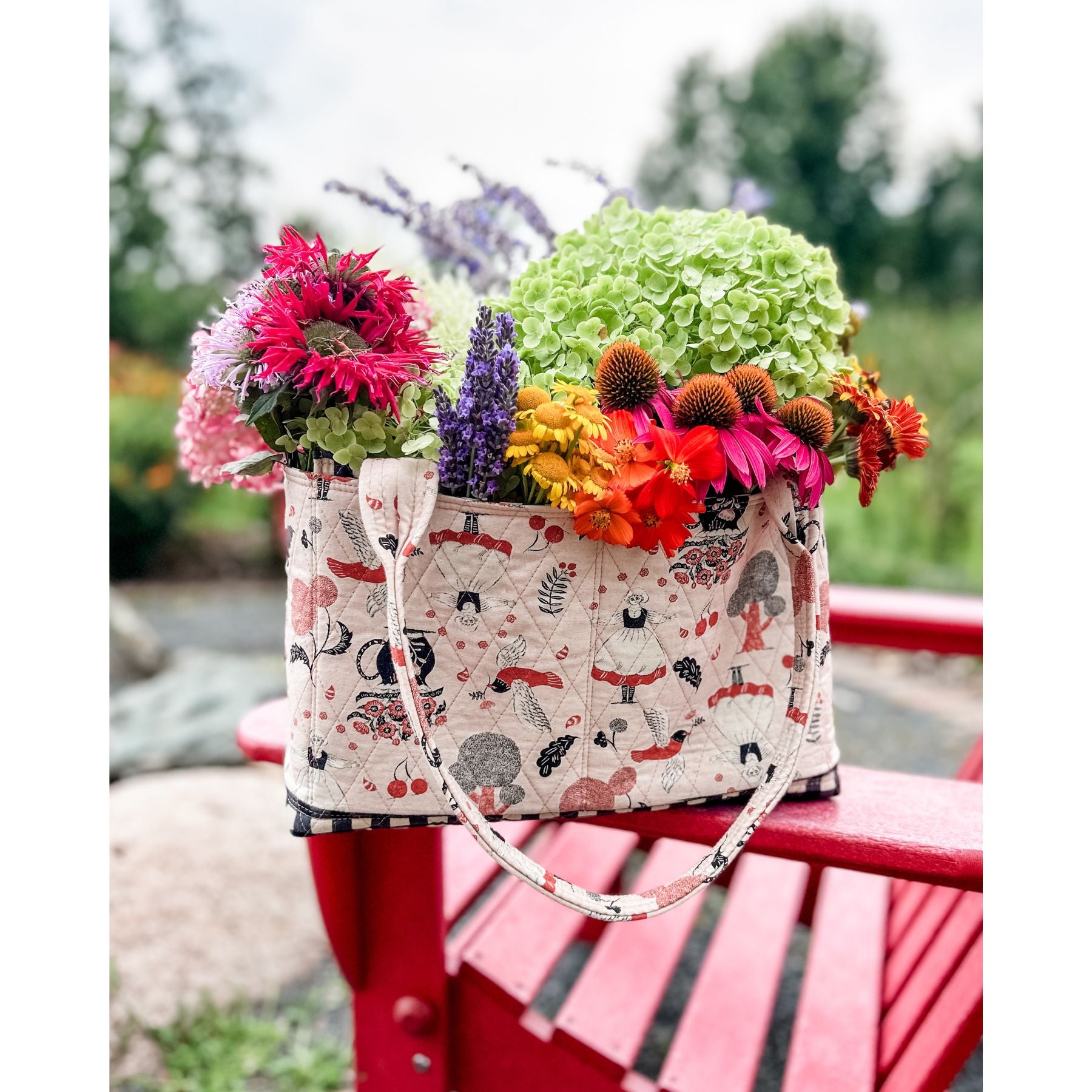 A Maggie Poppins Tote made with pink, red, black and white fabric filled with a garden bouquet, sitting on a red adirondack chair.