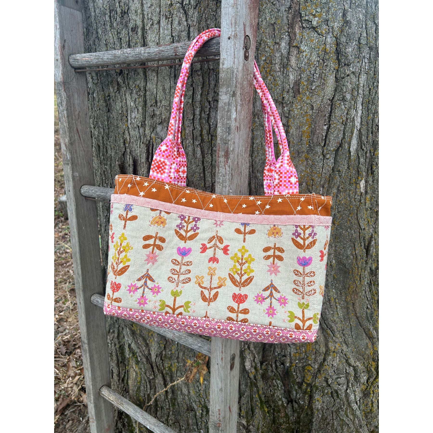 A Maggie Poppins Tote made in pink, brown, purple, yellow with floral fabric and embroidery detailing, hanging on a vintage ladder against a tree.