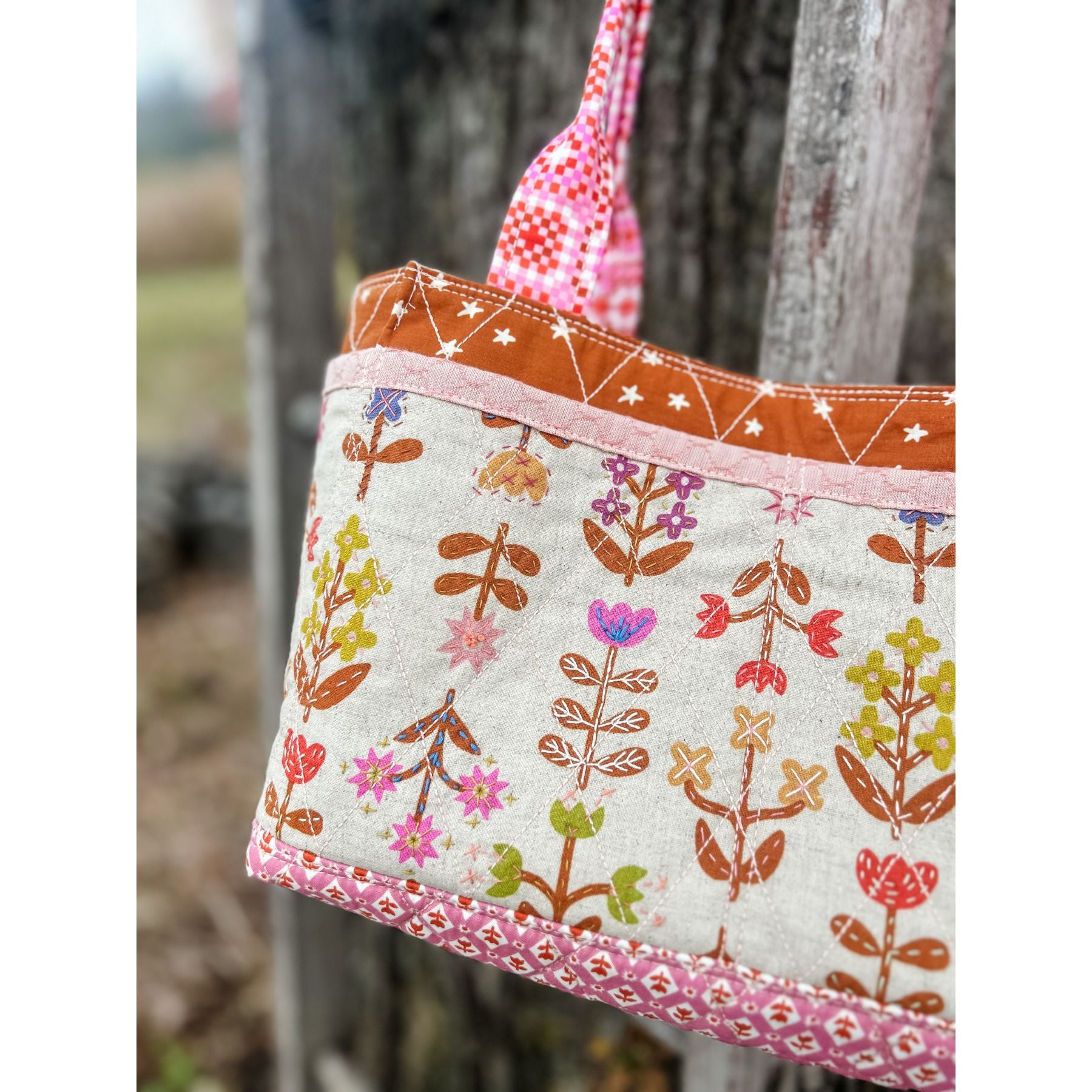 A close up of the a Maggie Poppins Tote made in pink, brown, purple, yellow with floral fabric and embroidery detailing, hanging on a vintage ladder against a tree.