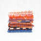 A stack of folded patterned fabric pieces in various colors and designs, including orange, pink, blue, and white, arranged vertically on a clean white background.